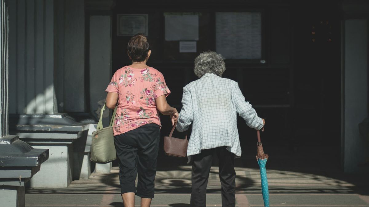 Zwei Frauen gehen, von hinten gesehen, eine Straße entlang; eine von ihnen stützt sich auf einen Gehstock.